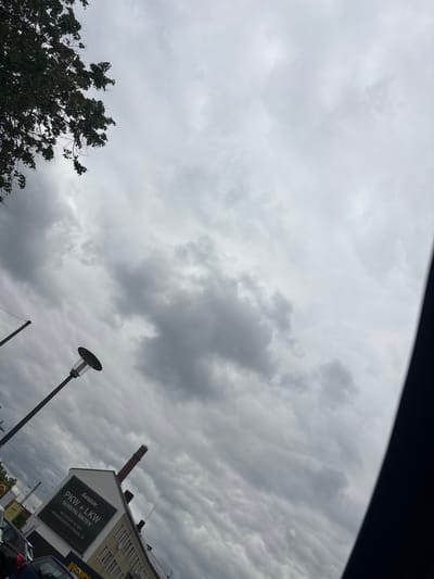 View of the sky and part of a street scene in Bielefeld, Germany. The sky is overcast with grey clouds. Below, there is a tree with green foliage on the left. A street lamp stands tall near the center of the frame. On the right, part of a building with a sign for Europcar PKW+LKW is visible. Cars are parked in the foreground.
