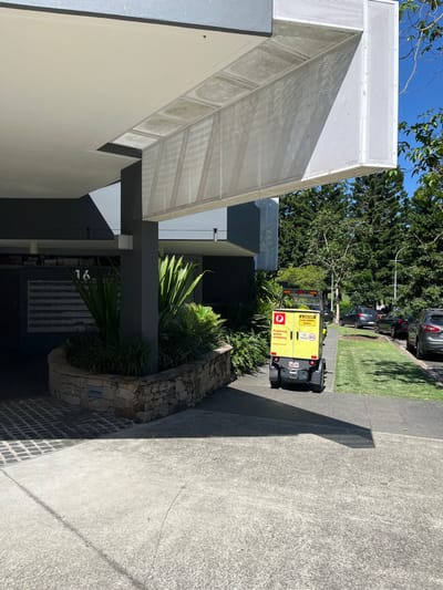 The image captures the exterior of a modern building entrance in Kelvin Grove, Australia, bathed in bright daylight under a clear blue sky. Distinct shadows are cast, suggesting it is mid-morning or afternoon.
The building exhibits a contemporary architectural style with light-colored walls and dark grey structural elements. A prominent feature is an angled, white perforated metal structure, possibly an awning or facade, extending over the entrance area. The building number "16" is visible near the entrance, alongside what appears to be a set of mailboxes or an intercom panel, indicating a multi-unit residential or commercial property.
In the foreground, a curved planter box constructed from natural, irregular stones holds a variety of green plants and foliage. The ground transitions from dark square pavers near the building to lighter, exposed aggregate concrete pathways.
Further down a paved driveway or path, a bright yellow delivery vehicle is visible from the rear. It is a compact, utility-style van or scooter, clearly marked as an Australia Post vehicle. A red rectangular logo on its back displays the text "Powering online shopping." Other visible text includes a yellow diamond-shaped sign stating "CAUTION TOP HEAVY VEHICLE," a "45 km/h" speed limit sticker, and the license plate "LZ 91Y." The presence of this vehicle suggests mail or parcel delivery activity.
Beyond the driveway, a strip of neatly mown green grass borders a street where several cars are parked. Numerous lush green trees form a natural backdrop against the bright sky. No people are visible in the scene.