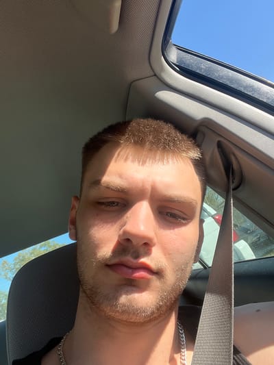 A close-up, front-facing image depicts a male individual seated inside a vehicle. The man has short, light brown hair and light facial stubble, wearing a dark garment and a silver-colored chain necklace. Strong sunlight illuminates the right side of his face, casting shadows. A beige seatbelt crosses diagonally from the upper right towards the lower right of the frame, secured to the vehicle's B-pillar. The car's interior features a gray headliner and pillar trim. A partially open sunroof is visible in the upper right, revealing a clear blue sky. Through the rear window, a portion of a light-colored vehicle and green foliage are discernible. The precise location within Jackson, United States of America, is not identifiable from the visual evidence.