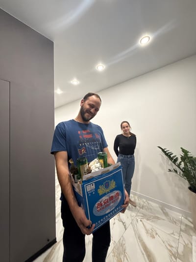 Two individuals are in a brightly lit interior space. A man in the foreground is carrying a case of Yanjing Beer. The woman in the background is smiling. The setting includes a white marble floor and a plant in a pot. There are recessed lights in the ceiling. The location is unknown and cannot be ascertained from the image.