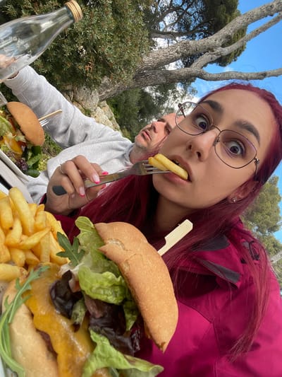 The image depicts an outdoor dining scene with two individuals at a table in Porto Cristo, Spain. In the foreground, a young adult female with long reddish-purple hair and glasses is holding a fork with a single french fry poised at her open mouth, looking directly at the camera with wide, expressive eyes. She wears a magenta outdoor jacket, and her left hand, visible, has pink french-tip nail polish and a ring on her ring finger. To her left, a white plate holds a mound of golden-brown french fries and a partially consumed burger topped with melted cheese, various lettuces, and a visible wooden skewer.
In the background, positioned behind the female and slightly to her left, an adult male wearing a grey hooded sweatshirt is seated. His head is tilted, and his eyes appear closed, suggesting he is resting or sleeping. A burger on a plate is visible in front of him. A clear glass bottle with a light-colored stopper stands on the table to the far left. The outdoor environment features dense green foliage from trees and bushes, a large tree with gnarled branches and sparse leaves, and a clear blue sky. Rocky terrain is visible in the distant background. The scene is illuminated by bright natural light, indicating daytime.