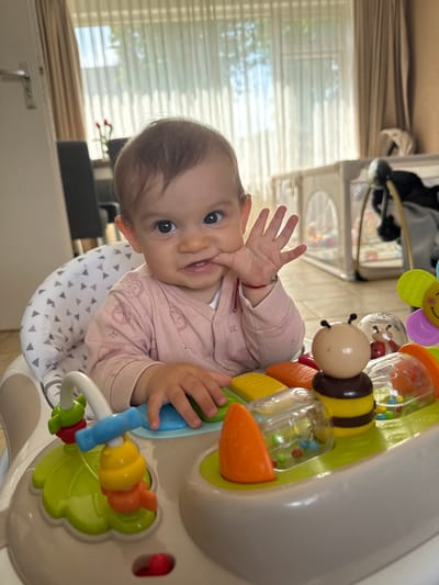 A baby sits in a white activity center with colorful toys, including a bee and a flower. The baby is wearing a pink sweater and has a hand near their mouth. The background shows a room interior with a window, curtains, a baby walker, and furniture. The image was taken indoors in Veghel, Netherlands.
