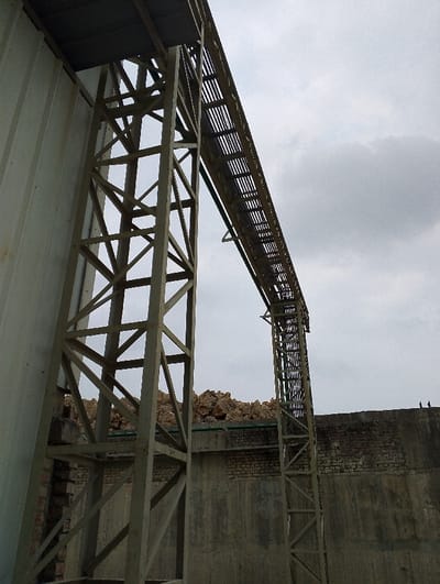The image shows a close-up view of a large metal structure located at 22.694, 90.601 in Pangasia, Bangladesh. The structure is composed of two large vertical beams connected by horizontal beams, creating a lattice-like pattern.  The structure appears to be a support for a long metal pipe system that runs above the beams, with the pipe system supported by a series of cross beams. There is a section of a white, corrugated metal wall behind the structure, with a portion of a brick wall visible in the distance. The sky is overcast.