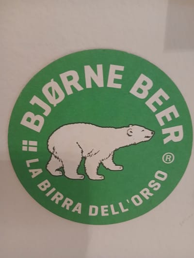 This is a circular green coaster with a white polar bear illustration in the center. The polar bear is depicted in a stylized manner, facing upwards with its mouth open, as if roaring or singing. The text on the coaster is in white, curved lettering. Around the top and right side of the bear, the text reads "BEER" and then continues in Italian to "DELL'ORSO". Along the bottom and left side, the text reads "LA BIRRA" and then "BJØRNE". There are also small graphic elements that appear to be the registered trademark symbol "®" near "BEER" and a double square symbol next to "LA BIRRA". The overall impression is that of a branded beer coaster, likely from a brewery named "Bjørne Dell'Orso" or similar, with the polar bear being its mascot. The setting is a close-up shot, possibly indoors, with a plain, light-colored background that appears to be a wall. There are no indications of time of day, weather, or specific emotional states conveyed by the image itself, beyond the dynamic pose of the polar bear. The location context of Monsummano Terme, Italy, suggests this coaster might be from a local establishment or a product distributed in that region.