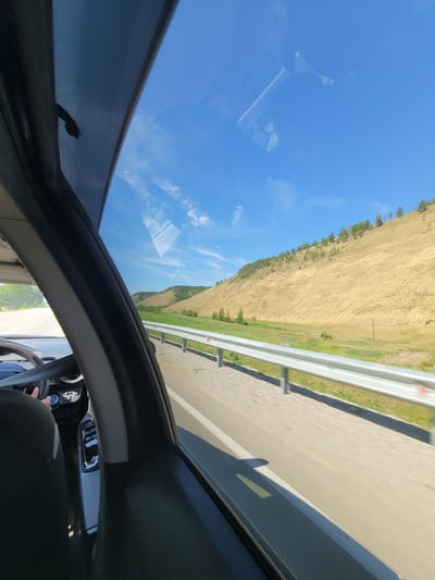 An outdoor, medium shot captures a perspective from inside a vehicle traveling on a road. The vehicle's interior is visible in the foreground, showing a steering wheel, a portion of the dashboard with a gear shift, and a seatbelt across the driver's chest. A person's hands are visible on the steering wheel, indicating they are driving.
The view through the vehicle's window reveals a vast landscape under a clear blue sky dotted with wispy clouds. To the right, a gently sloping hillside is covered in dry, golden grass and sparsely populated with coniferous trees. A metal guardrail separates the road from the hillside. The road itself is asphalt, with a white lane marker visible. The surrounding environment suggests a rural or semi-rural area, possibly in the city of Oktemtsy, Russia, given the context. The overall impression is of a scenic drive through a natural landscape on a sunny day.
