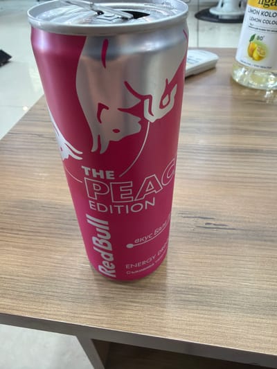 The image shows an open can of Red Bull The Peach Edition on a wooden surface. The can is silver and pink with the brand name and edition printed on the side. To the left of the can, partially visible, is a bottle of "LIMON KOLO/LEMON COLOGNE". The setting appears to be indoors, likely a table surface. The location, Dalgopol, Bulgaria, is inferred as the writing on the Red Bull can includes Bulgarian Cyrillic script.