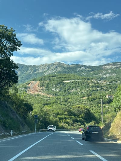 The image captures a view from a car driving on a paved road through a mountainous, forested area, possibly near Markovići, Montenegro. The road has clear white lane markings. On the right side of the road, a black Ford Focus with license plate "PG FT938" is in the foreground. Further ahead on the left, a white van is visible. In the background, a steep, tree-covered mountainside rises, with a section of exposed reddish-brown earth near the top. The sky is blue with scattered white clouds. A few utility poles are present along the roadside. The overall scene suggests a scenic drive through a rural, hilly region.