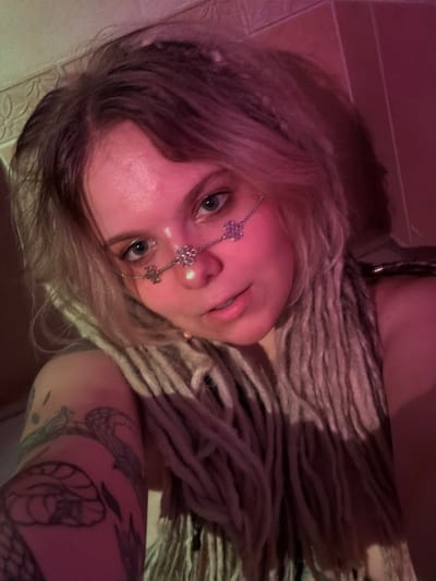 The media file shows a close-up, angled shot of a person with blonde dreadlocks adorned with a silver chain and flower-shaped jewelry on their nose. The person's eyes are a striking blue, and they have visible tattoos on their arm. The lighting is dim and casts a pinkish hue across the scene, suggesting it might be nighttime or indoors with colored lighting. The background appears to be a tiled surface and a textured wall, potentially in a home or studio setting. The overall mood is intimate and artistic, with the person's gaze directed towards the camera, conveying a sense of introspection or playful confidence. There is no visible text in the media file. The location context provided is Vostochny District, Russia, though this cannot be visually confirmed from the image alone.