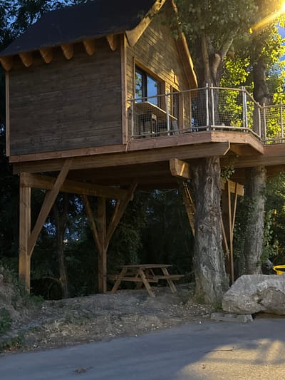 The image shows a treehouse constructed on thick tree trunks in the city of Grenay, France. The treehouse has a wooden exterior with a dark, sloped roof and a balcony with metal railings. A window is visible on the side of the treehouse. Below the treehouse, a picnic table is situated on a gravel-covered area. Large trees surround the structure, and a substantial rock is visible in the foreground. The scene is illuminated by artificial light, suggesting it is either dusk or nighttime.