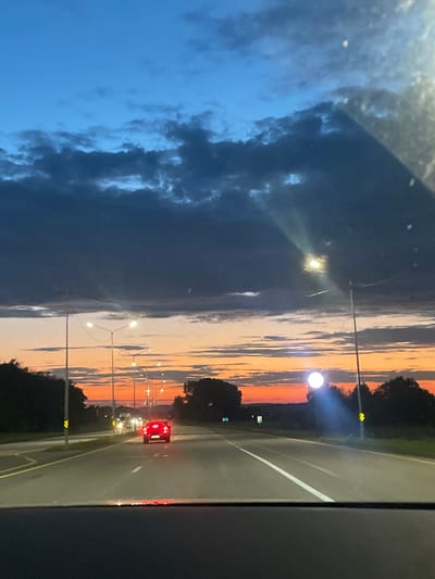 The image depicts a twilight scene on a highway, likely on the outskirts of Akzhibay, Kazakhstan. The sky is a gradient of deep blue at the top, transitioning to vibrant orange and pink hues near the horizon, suggesting the sun has recently set. Dark, dramatic clouds are visible against the lighter sky. A paved highway stretches into the distance, with multiple lanes marked by white lines. Streetlights, illuminated, line the right side of the road, casting light on the asphalt. In the middle of the highway, a red car is driving away from the viewer, its taillights illuminated. Trees and vegetation flank the sides of the road, with more distant hills visible on the horizon. The perspective suggests the image was taken from inside a vehicle, looking forward through the windshield, with a portion of the vehicle's dashboard visible in the foreground.