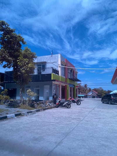 In Aluepiet, Indonesia, a scene unfolds under a vast, bright blue sky adorned with wispy white clouds. The foreground is dominated by a grey, concrete-paved area, likely a street or parking lot, with visible expansion joints and some debris scattered on the ground. To the left, a multi-story building displays a vibrant facade with sections painted in red, pink, green, and grey. The building features balconies, windows, and awnings. A mature tree with dense green foliage stands tall beside the building, its branches extending towards the sky, and a string of small, bare lightbulbs is visible hanging from its lower branches. A low wall constructed of various-sized rocks borders the area in front of the building. In front of the building, parked on the concrete, are two motorcycles: one is red and white, and the other is red. Further into the background, a black car is parked on the right side of the paved area. Beyond the building, residential houses with red roofs and a gas station canopy are visible, interspersed with palm trees and other greenery, suggesting a mixed urban and residential environment.
