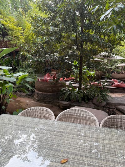 The image shows an outdoor seating area at a cafe or restaurant in Tambon Mae Raem, Thailand. In the foreground, a glass-topped table with a wicker base reflects the surrounding greenery and sky. Three wicker chairs are visible behind the table. The middle ground features a natural landscape with lush vegetation, large rocks, and pathways. Several seating arrangements are scattered throughout the scene, including low wicker tables with large, round cushions in a coral color, and larger, woven basket-like seating structures filled with similar cushions. In the background, trees and foliage create a dense canopy, with glimpses of umbrellas and possibly other seating areas. The overall atmosphere is relaxed and natural, typical of an outdoor dining establishment in a tropical setting.