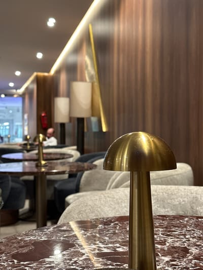 The image captures the interior of a modern, upscale restaurant or lounge, characterized by warm lighting and rich materials. The perspective is from a low angle, looking across a row of tables and seating arrangements.

In the foreground, a polished brass or gold-colored lamp fixture with a semi-circular shade extends upwards from a vertical brass rod. To its right, a dark red marbled tabletop, streaked with white veins, occupies the bottom right corner. Behind the table, a light grey upholstered chair or booth seating is visible.

A series of similar brass lamp fixtures, marbled tables, and grey upholstered seats recede into the mid-ground, creating a symmetrical arrangement. Several blurred figures are seated at these tables, suggesting an active dining or social scene. One figure in the mid-background, wearing what appears to be a light-colored top, might be a staff member. A small, blurry red object rests on one of the tables further back.

The background features a dark wall on the left, illuminated by a continuous strip of warm LED lighting. Several circular, out-of-focus lights (bokeh) are scattered on this wall. The ceiling or upper wall above the tables is covered in dark wood panels with a distinct horizontal grain. In the far background, a large, blurry area with bright reflections suggests a screen or window, but no specific details or readable text are discernible.

The overall atmosphere is warm and sophisticated, indicative of an evening setting due to the prominent artificial lighting. The dominant materials—polished brass, dark wood, and red marble—contribute to the establishment's luxurious aesthetic. No specific text is visible in the image.