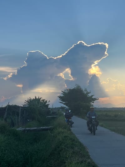 This media file depicts a scenic view of a rural landscape in Đà Nẵng, Vietnam, during what appears to be late afternoon or early evening. The primary subjects are two people riding motorcycles on a paved road that winds through lush green fields and vegetation. The sky is dramatic, dominated by large, billowy clouds illuminated by the setting sun, which creates a striking contrast between dark, shadowed areas and brightly lit edges. Rays of sunlight pierce through the clouds, casting a warm, golden glow. One motorcycle rider is closer to the foreground and more in focus, while the second is further down the road. The setting is characterized by verdant grass and trees lining the road, with distant hills or mountains faintly visible on the horizon, suggesting a rural or semi-rural environment. The overall mood is peaceful and serene, enhanced by the beautiful play of light and shadow in the sky. No text is visible in the image.