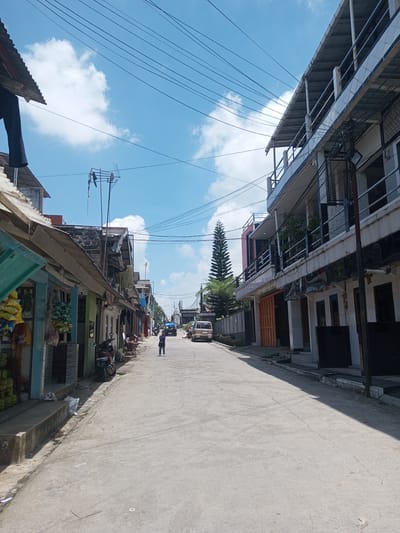 A street in Kioumaknao 2, Laos, presents an asymmetric urban landscape. The left side is characterized by a row of older, more modest structures, possibly shops or small businesses, indicated by displayed goods, awnings, and a motorcycle parked in front. Several people sit and interact.
The right side features taller, more modern buildings with multiple stories and balconies. A vehicle is parked further down the street, and a single person walks in the middle of the road. Power lines stretch across the blue sky with scattered clouds, adding to the urban context. The road surface is concrete.