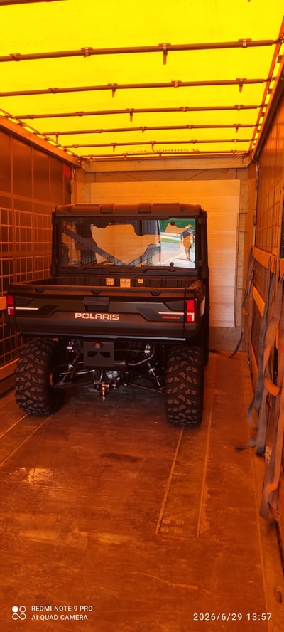 An all-terrain vehicle (ATV) is parked inside a covered trailer or truck. The ATV is black and has the brand name "POLARIS" visible on the rear. The trailer's interior is illuminated by a yellow tarp that covers the roof and is supported by metal rods. The floor of the trailer appears to be made of wood or metal and shows some wear and tear. Straps are visible on the right side of the trailer, suggesting the ATV may have been secured. The image has a timestamp in the bottom right corner indicating June 29, 2026, at 13:57. There are no people visible in the immediate vicinity of the ATV or within the trailer. The scene is indoors and appears to be a storage or transport situation for the ATV.