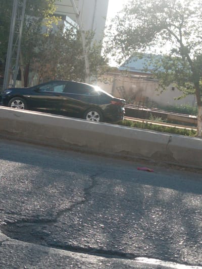In Bagat, Uzbekistan, a black sedan is parked on a paved road. The car, facing left, is a compact four-door model with tinted windows. Its front wheel is turned slightly towards the left. The vehicle is positioned next to a raised concrete barrier, which runs parallel to the road. Beyond the barrier, a lush green embankment with various plants and trees is visible. In the background, several buildings can be seen. One structure appears to be a modern building with a reflective facade, possibly a hotel or office building, while another has a blue roof and beige walls. Sunlight filters through the trees, casting a warm glow on the scene and creating lens flare effects. The road itself shows signs of wear, with visible cracks and uneven surfaces. A small pink object, possibly a piece of litter, lies on the asphalt near the concrete barrier. The overall impression is a sunny day in an urban or semi-urban setting.