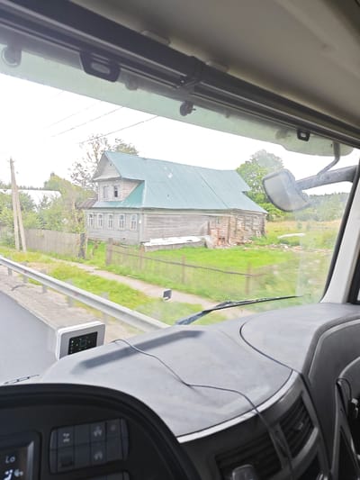 The image is taken from inside a vehicle, likely a truck, looking out the windshield. In the foreground, the dashboard is visible, with some control buttons and a small electronic device mounted on the windshield. The scene outside the vehicle shows a rural landscape. A wooden house with a green roof is prominent, situated behind a wooden fence. There is green grass and trees surrounding the house. A metal guardrail runs along the side of a paved road in the midground. The sky appears overcast. The location is identified as Golenikha, Russia.