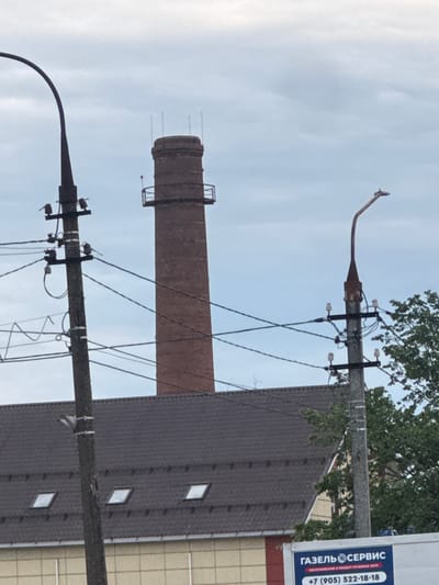 The image shows a view of a brick smokestack in the city of Dedovsk, Russia, on a cloudy day. In the foreground, a dark, curved lamppost stands to the left. To the right of the smokestack, another lamppost with a curved arm is visible. Several utility poles with electrical wires crisscross the scene. Beneath the smokestack and lampposts is the dark, sloped roof of a building with several small, square skylights. The building's facade, visible below the roofline, is light-colored with a grid pattern. A portion of a white truck or van with a blue and red sign featuring Russian text and a phone number is present in the bottom right corner. The environment appears to be urban or industrial.