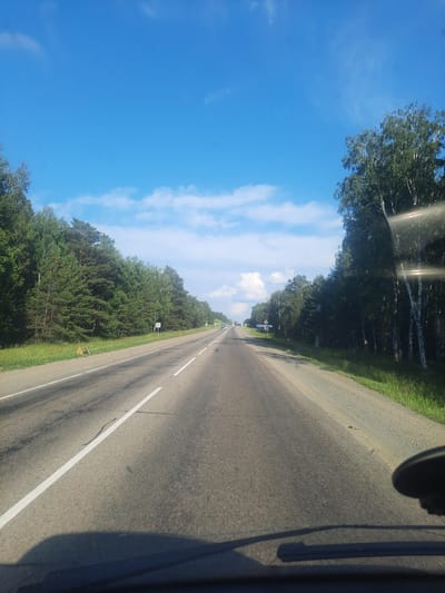 The media file captures a view from inside a vehicle traveling on a paved road through a rural landscape. The road stretches into the distance, marked by white lines. On the left side of the road, a dense forest with a variety of trees borders the asphalt. The sky is a clear blue with scattered white clouds. Sunlight creates reflections and glare on the windshield of the vehicle from which the photo is taken, casting shadows. In the distance, a few other vehicles can be seen on the road, suggesting it is in use. The time of day appears to be daytime, with bright sunlight. The overall impression is of a journey through a natural, serene environment. There is no visible text in the image. The location context provided is Баюновские Ключи, Russia.