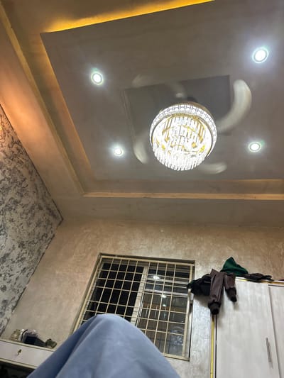 The image displays an interior room, likely a residential space, in Yalwa, Nigeria, viewed from a low-angle perspective. The ceiling features a modern design with a central, multi-tiered chandelier adorned with numerous clear, crystal-like elements and a gold-toned frame. A ceiling fan with blurred blades, indicating motion, is integrated into the chandelier fixture. Four circular recessed spotlights emit bright white light from the light grey ceiling, which also includes a yellow-orange cove lighting strip along its perimeter. The left wall exhibits a textured, mottled grey and white pattern. The adjacent wall beneath the ceiling appears to be a plain, light grey-beige with a slightly coarse finish. A rectangular window, secured by a light-colored metal griller in a grid pattern, is visible towards the bottom right, with internal light reflections on its panes. A light-colored, two-door wardrobe with silver horizontal handles is partially visible on the right, topped by a draped dark green garment and dark brown trousers. On the far left, a light-colored surface holds a white jar and another dark object. The immediate foreground is occupied by light blue fabric, obscuring the lower portion of the scene. The room is actively illuminated by artificial lighting.