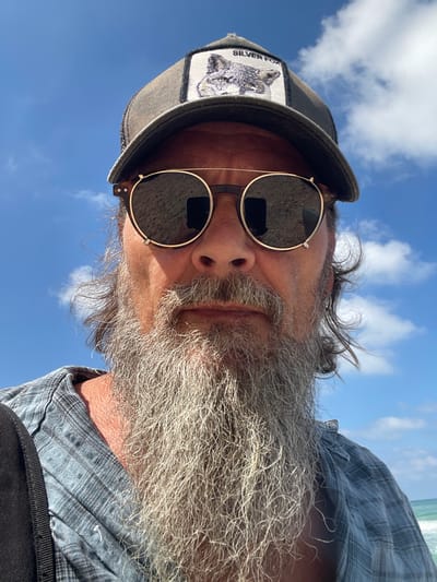 A man with a long, gray beard and a blue plaid shirt is seen from the chest up, taking a selfie. He is wearing a brown trucker hat with a patch that says "SILVER FOX" and a picture of a fox. He also has round, gold-rimmed sunglasses with dark lenses. The background is a bright blue sky with scattered white clouds, and a hint of the ocean can be seen on the right side of the image, suggesting the location is a beach. The man's skin appears sun-kissed. The lighting indicates it is daytime. There is a black strap visible on the left side of the frame, possibly from a bag.