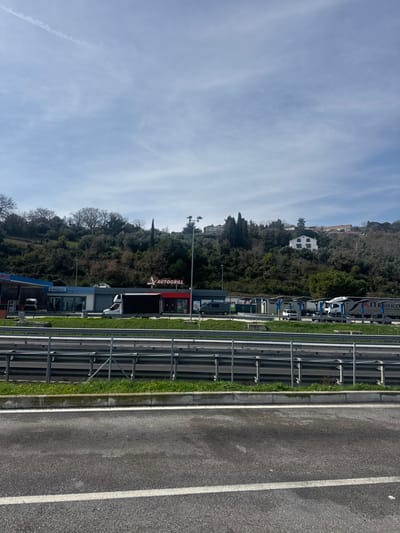 The image displays a roadside service area in Tortoreto, Italy, observed from a low angle across a multi-lane highway. In the foreground, a dark asphalt road with a solid white line on its right edge transitions into a grassy median and a three-tiered metal guardrail system.

The midground features a commercial service station complex. A central building has a prominent red section displaying "AUTOGRILL" in white lettering. To the left, a blue-canopied fuel station is visible. A white truck with a dark enclosed trailer is parked directly in front of the Autogrill building, while a grey van is parked to its right. Further right, a dedicated parking area accommodates multiple large commercial trucks and semi-trailers, one bearing the "MASSUCCI" brand. Tall, multi-headed streetlights illuminate the service area.

The background is dominated by a densely vegetated hillside covered with a mix of evergreen and deciduous trees. Several structures are discernible on the slope, including a distinct white two-story house with a reddish roof on the upper right side of the hill. The sky is light blue with sparse, wispy white clouds, and a single aircraft contrail is visible in the upper left quadrant. No individuals are clearly identifiable or engaged in specific actions.