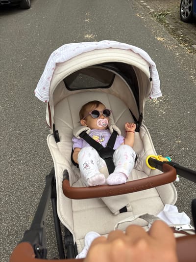 This media file shows a baby lying in a stroller on a paved surface that appears to be a sidewalk or road. The baby is wearing a purple t-shirt with Winnie the Pooh characters and patterned pants. Pink sunglasses are on the baby's face, and a pink pacifier is in its mouth. A red string bracelet is on the baby's left wrist. The stroller is light beige with a brown handle that has the embossed text "life is a miracle". A white and pink patterned blanket is draped over the edge of the stroller. A yellow toy ring is attached to the stroller's handle. In the background, the wheel of a blue car is visible on the left side of the image. The scene suggests an outdoor setting, possibly a street or park in Veghel, Netherlands. The lighting indicates it is daytime. The overall mood is peaceful, with the baby appearing relaxed in the stroller.