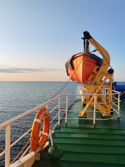 A perspective shot from the deck of a ship shows an orange life raft secured by a yellow davit crane against a pale blue sky. The calm, dark blue ocean stretches to the horizon, meeting a soft band of orange and pink sky. A white railing runs along the edge of the green deck, with an orange life preserver hanging from it. A small, green, spherical object with a valve sits on the deck near the life preserver. The ship appears to be in open water, likely at dawn or dusk, given the lighting. There are no people visible in the frame. The text on the life raft is in Cyrillic and appears to read "Евгений Орловский" and "КОТЛАС".