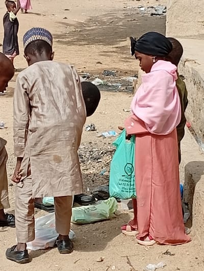 Several children stand outdoors amidst litter. One boy wears a tan outfit with a patterned cap, his back to the camera. A girl beside him is dressed in a long peach gown and a pink headscarf. She holds a green plastic bag. Another boy stands slightly behind her. More children and scattered debris are visible in the background. The ground is sandy and uneven. A wall structure is present on the right.