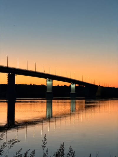The image depicts a bridge over a body of water at dusk. The sky exhibits a gradient of colors, transitioning from a deep blue at the top to a vibrant orange near the horizon. The bridge, silhouetted against the colorful sky, is illuminated by streetlights along its length. Two large concrete pillars support the bridge, with their reflections visible in the calm water below. A dark line of trees forms a silhouette along the far bank of the water. In the foreground, the edges of some dry grasses are visible, adding a touch of natural detail to the scene. The location is identified as Tarasovo, Russia.