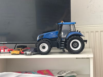 The media file shows a collection of toy vehicles, including a large blue tractor and several smaller cars, displayed on a white shelf. The tractor is a prominent object, positioned on the right side of the frame, with its large wheels and detailed cabin visible. To the left of the tractor, a row of toy cars is lined up. These include a red car with the number 7, a yellow sports car, a red sports car, and a silver and blue sports car. The toys are placed in front of a dark screen, likely a television or monitor, which dominates the background. To the right of the screen, a white wall is visible, with some scribbled drawings on it, possibly made by a child. A radiator is also visible below the wall. The lighting suggests an indoor setting, likely a room. The overall scene depicts a display of children's toys, with no specific activity taking place beyond their arrangement. The image is taken from a slightly low angle, looking up at the shelf. There is some text visible on the dark screen that reads "KrügeraMatz".