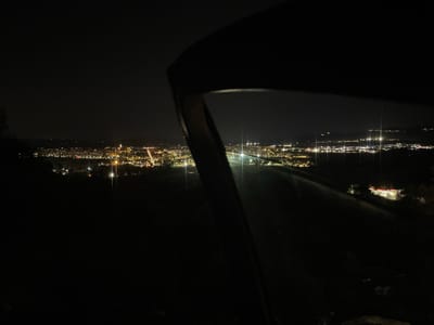 The image presents a wide-angle, elevated view of Pacs del Penedès, Spain, at night. The scene is dominated by the sprawling lights of the town and surrounding areas, which stretch across the middle ground and into the distant horizon. The sky above is dark, creating a stark contrast with the numerous yellow, orange, and white artificial lights emanating from buildings, streetlights, and possibly vehicles.

In the immediate foreground, a large, dark, curved object occupies the right side of the frame, partially obscuring the view. This object appears to be a structural element, possibly a window frame, a railing, or a car's door frame, and shows subtle reflections on its surface.

The town itself is a tapestry of light, with a more concentrated cluster of illumination in the center-left. A distinct, brighter yellowish-orange light source, possibly a church or a significant building, is noticeable within this cluster. Further to the right, in the mid-ground, a brightly lit commercial building stands out with prominent red and white lights, suggesting a gas station or a similar establishment.

The time of day is clearly night, with no natural light sources visible. The weather appears to be clear, allowing for crisp visibility of the distant lights without significant atmospheric interference. No people or legible text are visible in the photograph. The scene evokes a sense of calm observation of a lively nocturnal landscape.