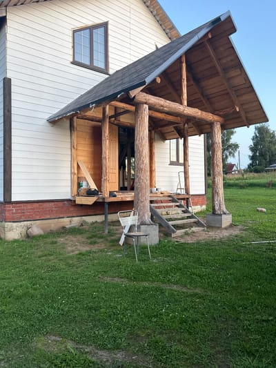 This image depicts the exterior of a house under construction in Vereiki, Russia. The house is clad in white horizontal siding with a visible red brick foundation. A wooden porch with a dark shingled roof is a prominent feature. The porch is supported by thick, natural logs, giving it a rustic appearance. Wooden stairs lead up to the porch, and a ladder is visible leaning against the house. Various construction materials and tools, including cans of paint and wooden planks, are scattered on and around the porch. The surrounding environment is a grassy yard with some trees in the background. The sky is clear blue, suggesting fair weather. No people are visible in the image.