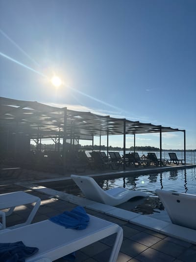 The image depicts an outdoor swimming pool area with lounge chairs and a covered seating structure, situated by a body of water, likely a river or lake, with a tree-lined shore visible in the distance. The sun is bright in the upper left quadrant of the sky, casting lens flare. The pool features an infinity edge and some lounge chairs are partially submerged in it. Blue towels are draped over lounge chairs on the paved poolside deck in the foreground. The scene is set in Kamiani Potoky, Ukraine. There are no visible people in the immediate foreground or middle ground, though one person appears to be lying on a lounge chair under the covered structure in the middle ground.