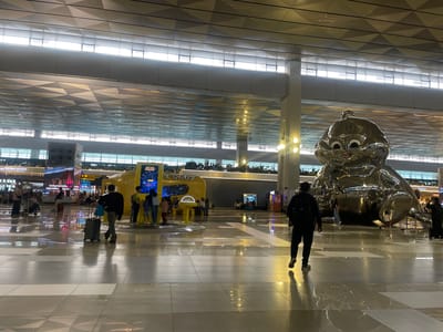 The image depicts the interior of an airport terminal. In the foreground, a man in a black jacket and pants walks away from the camera, pulling a rolling suitcase. The floor is polished tile with a reflective surface, creating mirrored images of the ceiling and lights. In the mid-ground, a large, shiny, silver sculpture resembling a cartoon character is prominently displayed. To the left of the sculpture is a yellow, spaceship-like structure with a display screen and several people gathered around it. The background shows more of the airport terminal, including signage, shops, and other travelers. The ceiling features an intricate geometric design with integrated lighting. Based on the visible signage indicating "Soekarno-Hatta International Airport," the location is likely within the city of Tangerang, which is part of the Jakarta metropolitan area, not Pajang, Indonesia.