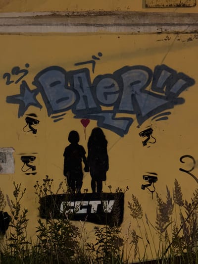 The image is a photograph of graffiti on a yellow wall, likely in the city of Imeni Sverdlova, Russia. The graffiti includes large blue lettering, a silhouette of two children holding hands, a small red heart above them, and several stenciled images of security cameras. Below the figures, a black rectangular tag with white lettering reads "CCTV." The wall is weathered and shows signs of age. In the foreground, tall dry grass and plants obscure the bottom portion of the wall and the graffiti. The lighting suggests it may be nighttime or a dimly lit area.