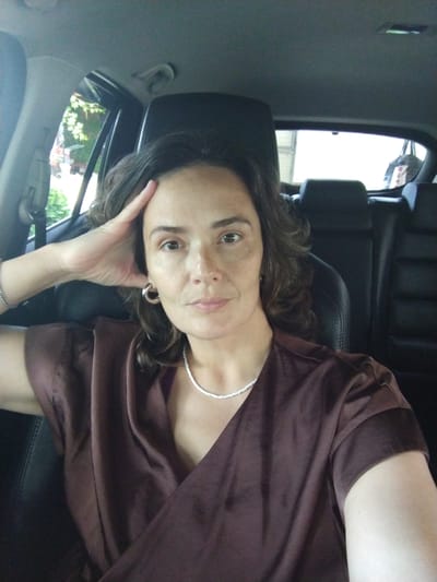 A woman is sitting in the driver's seat of a car, with her left arm resting on her forehead. She is wearing a dark brown satin top and a silver beaded necklace. The interior of the car, including black leather seats and headrests, is visible behind her. Through the car windows, greenery and trees are visible, suggesting an outdoor location. The scene appears to be a portrait taken inside a vehicle. The exact location within the city of Vinogradny, Russia cannot be determined from the image alone.