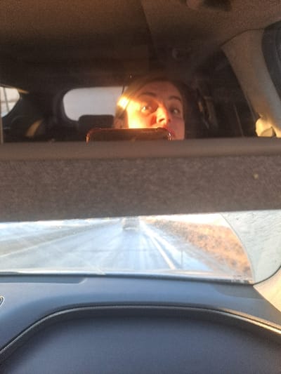 The image is a vertical composition, appearing to be taken from inside a vehicle, possibly a car, with the primary subject being a reflection of a person in a glass surface.

In the left half of the image, a reflection of a person's face is clearly visible, positioned upside down. The person has light-colored hair, and their eyes are open and looking directly towards the viewer. A strong, bright light illuminates the lower part of their face (when upright), specifically the nose and cheek area, creating a warm, golden glow. A smartphone or similar device, held by the person, is also reflected, partially obscuring the mouth area. The back of the phone case appears to be a reddish-brown color. The background behind the reflected face is dark and indistinct, suggesting the interior of the vehicle.

To the right of the reflected face, what appears to be part of the car's interior is visible, possibly a dark blue or gray door panel. Further to the right and occupying the right half of the image, there is a bright outdoor scene viewed through a window or windshield. This view is somewhat blurred and distorted by the glass surface, showing abstract patterns of light and shadow, possibly suggesting trees or buildings passing by, or simply the textured surface of the window itself. Streaks of bright light cut across this area, indicating strong sunlight.

The overall impression is of a sunny daytime scene, with sunlight intensely reflecting off surfaces. The person seems to be looking at their own reflection, possibly taking a selfie, given the position of the phone. No text is visible in the image.