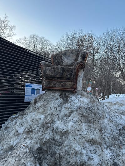 An ornate, dark-patterned upholstered armchair, exhibiting visible wear and featuring dark wood scrollwork trim, is positioned atop a substantial mound of dirty, melting snow in Sadovaya, Russia. To the left of the snow pile, a large, dark, horizontal slatted structure runs diagonally upwards, with a white public information sign displaying Cyrillic text and illustrations of recycling bins attached near its base. Bare deciduous trees with thin branches against a clear, light blue sky dominate the upper background. Partially obscured by snow and trees in the right background are glimpses of colorful, decorative objects. No individuals are present in the scene.