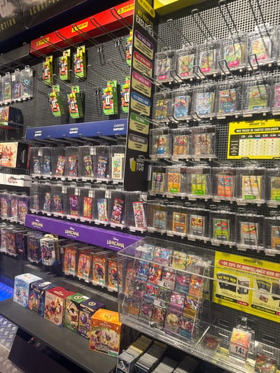 The image shows a retail store in Leers, France, with a focus on collectible trading cards. The store displays a wide variety of trading card game products, including booster packs, boxes, and individual cards, organized on shelves and in clear display cases. Prominent brands visible include Pokémon, Lorcana, and Avatar. Promotional signage for "Univers TCG" is present, highlighting exclusive card packs and offers, including a "20% de remise avec nos kits" (20% discount with our kits) and a "Kit Collectionneur" (Collector's Kit). The products are arranged neatly, with pricing information visible on some of the displays. The environment appears to be an indoor retail setting, likely a specialized hobby or game store.