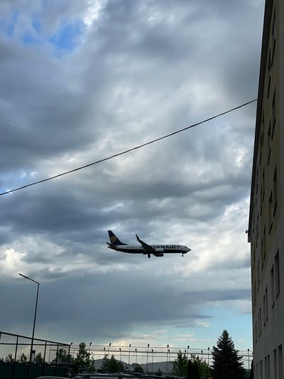 The image depicts a Ryanair airplane taking off from Otopeni, Romania, viewed from below, with a cloudy sky in the background. An apartment building is visible at the top of the frame, and a fence, trees, and streetlights are seen near a parking lot. A power line stretches across the sky.