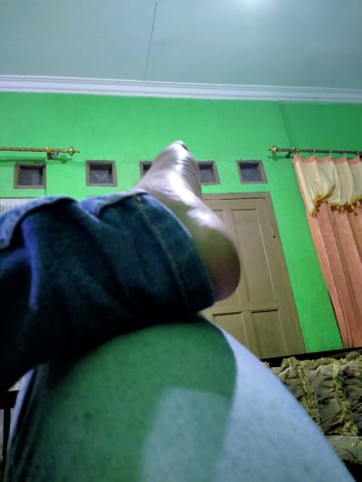A person lies down with their right foot resting on their left thigh. They wear blue jeans and a light gray t-shirt. Behind them, a green wall has a brown door, four small ventilation squares, and a curtain. The ceiling is white, and the room has a dim, possibly artificial, lighting. Bundles of jute or similar natural fiber are visible in the lower right corner.