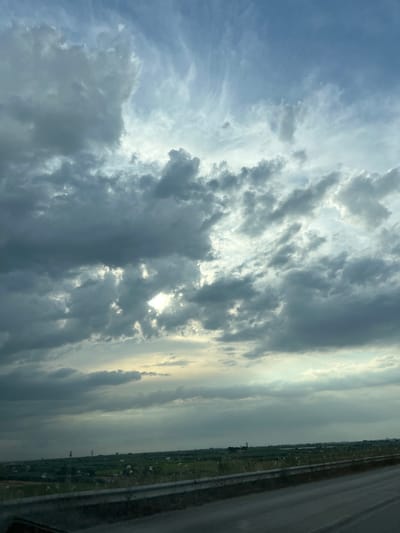 The image displays a panoramic view of the sky and landscape, likely taken from a moving vehicle on a road. The sky is filled with dramatic, overcast clouds, ranging in color from dark grey to lighter shades, with some patches of sunlight breaking through. The clouds appear dense and textured, suggesting impending or recent rainfall. Below the sky, a flat, green expanse of land stretches to the horizon. The landscape is dotted with what appear to be agricultural fields, some with visible white objects, possibly livestock or scattered vegetation. In the distance, several tall, slender structures, resembling utility poles or small towers, are visible. A concrete barrier runs along the edge of the road in the foreground, with a paved surface, presumably a highway or road, occupying the bottom portion of the image. The overall impression is of a rural or semi-rural setting under a moody, overcast sky. The location is identified as Barletta, Italy.