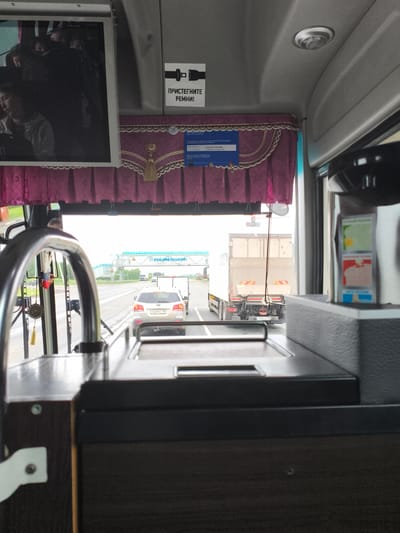 The image depicts the interior of a bus, viewed from the driver's or front passenger's perspective, looking forward through the windshield onto a highway. The scene is set in the city of Ижевск, Russia, as indicated by the "18" regional code on visible license plates, corresponding to the Udmurt Republic where Ижевск is the capital.
Inside the bus, a dark-colored dashboard or console is visible in the foreground. A chrome handle or railing curves across the upper left. Directly above the windshield, pink and gold patterned curtains with decorative tassels are drawn across a pelmet. A blue information sheet, possibly a license or route details, is attached to the curtain rail, partially visible with text including "ООО", "ЛИЦЕНЗИЯ", and dates "01.08.2016 по 01.05.2017". To the right of the curtains, a white rectangular sign with a black seatbelt icon reads "ПРИСТЕГНИТЕ РЕМНИ!" (Fasten your seatbelts!). An overhead monitor on the left displays the blurred image of several people, one appearing to be a young person or child looking down. On the far right, a beverage dispenser or coffee machine is partially visible, featuring colorful labels with Russian text such as "ПЕРВЫЙ РЕЖИМ", "ВТОРОЙ РЕЖИМ", and "ТРЕТИЙ РЕЖИМ" (First Mode, Second Mode, Third Mode). The bus ceiling is light gray with a recessed light fixture.
Outside, the bus is on a multi-lane highway under an overcast sky. Traffic flows in the same direction. Immediately ahead, a silver Kia Sorento SUV (license plate T469MY 18) is followed by a white trailer. Further ahead, a white semi-truck with a trailer (license plate T316TC 18) is visible. In the distance, an overhead gantry structure spans the highway, displaying a large blue sign with the text "РОСАВТОДОР" (ROSAVTODOR - Russian Federal Highway Agency) in white Cyrillic letters. Green foliage and trees line the sides of the highway.