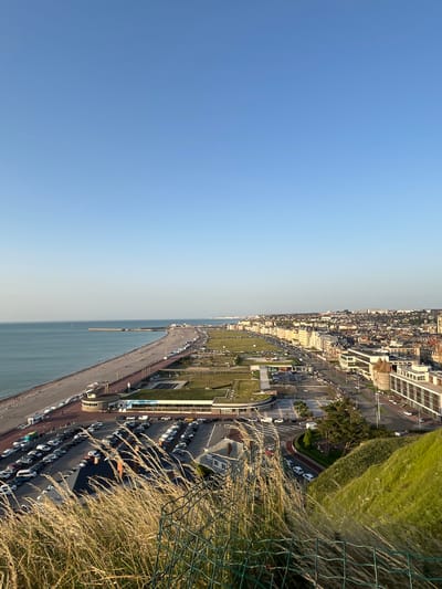This media file captures a high-angle view of a coastal cityscape, likely Dieppe, France, with a clear blue sky dominating the left half of the frame. The right half of the image reveals a well-developed urban coastline, featuring a long, curving beach or promenade that meets the calm, blue sea. Beyond the promenade, a street lined with parked cars runs parallel to the coast, interspersed with patches of green parkland and trees.
Buildings of various sizes and architectural styles are visible along the street, suggesting a populated area. The architecture appears to be a mix of modern and older structures, with some buildings exhibiting a classic European aesthetic. A large parking lot filled with numerous cars is a prominent feature, indicating a popular destination, likely for beachgoers or tourists.
In the foreground, dry, wispy grass and a wire fence are visible, suggesting the viewpoint is from a slightly elevated position, perhaps a cliff or a tall building overlooking the scene. The lighting indicates it is late afternoon or early evening, with the sun casting a warm glow over the scene, highlighting the textures of the grass and the architectural details. The overall mood is peaceful and serene, characteristic of a pleasant day at the coast. No text is visible in the image.