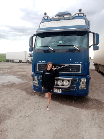 A young woman stands in front of a large blue Volvo truck. She is wearing a black hoodie and black shorts, and her right hand is raised in a peace sign. The truck is parked on a gravel surface, with a large white semi-trailer truck visible in the background. The sky is overcast, suggesting a cloudy day. The license plate on the Volvo truck reads "KG 545 01 AJY." Text visible on the truck includes "VOLVO," "FH 16," and "KING OF THE ROAD." The scene appears to be a truck stop or parking area, possibly in Sarytogay, Kazakhstan, given the license plate and the overall landscape. The young woman's pose and facial expression suggest a casual and cheerful demeanor. The setting is outdoors with dirt and scattered puddles indicating potentially wet conditions.
