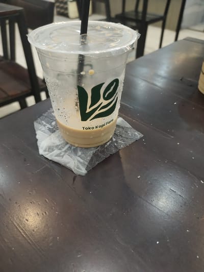 A close-up, eye-level shot shows a disposable plastic cup containing a light brown beverage, possibly coffee with milk, with a straw inserted. The cup is resting on a crumpled, translucent napkin placed on a dark wooden table. Condensation is visible on the exterior of the cup, with a few drops of liquid on the napkin. A green logo with the text "Toko Kopi Fora" is printed on the cup. The background is blurred, showing dark chairs and tables, suggesting an indoor seating area of a cafe or restaurant. The overall lighting is dim, with some reflections on the table surface. This scene appears to be in Lhoksukon, Indonesia, given the context.
