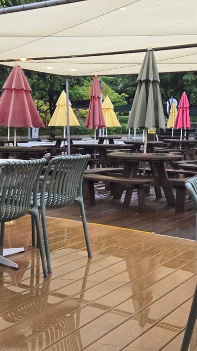 The image depicts an outdoor seating area, likely a cafe or restaurant, under a large, cream-colored awning. The scene appears to be in Gwangmyeong, South Korea, as indicated by Korean signage visible on a post in the background. The area is furnished with numerous wooden picnic tables and benches, many of which are occupied by closed umbrellas of various colors, including pink, yellow, and green. The wooden deck flooring is wet, with visible reflections, suggesting it has recently rained or is still raining. In the foreground, two grey metal chairs with a slatted design are positioned on the wet deck. The overall atmosphere is subdued, likely due to the weather, with no people actively engaged in the space. Lush green trees form a backdrop behind the seating area.