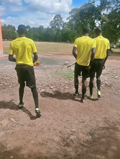 In Lwandeti ward, Kenya, three adult males, dressed in yellow short-sleeved shirts with black trim, black shorts, black socks, and athletic cleats, stand with their backs to the observer on a reddish-brown dirt and gravel surface. The individual on the far left wears black compression tights and holds a red and yellow checkered flag. The central figure among the two on the right holds a dark stick-like object. Their collective attire suggests roles as sports officials. They face a large, light brown grassy field, bordered by a dense line of trees under a partly cloudy blue sky. A light-colored building or wall structure is partially visible on the far left. Distant figures are scattered across the field, and a motorcycle is partially visible on the far right. Shadows are cast forward, indicating bright daylight.